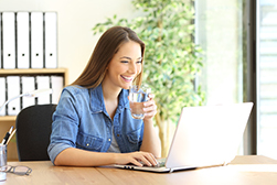 Young woman with straight long brown hair drinking water at her desk.