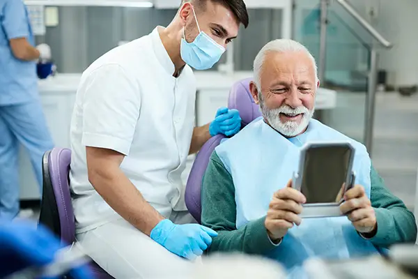 Happy, senior patient looking at himself in a mirror with dental implants at Lakewood Dental Arts in Lakewood, CA